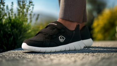 Close-up of feet in black Giesswein trainers with white soles on a gravel path. The person is wearing brown trousers and green plants and mountains can be seen in the blurred background.