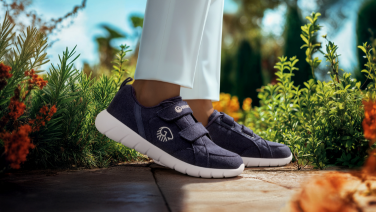 Close-up of feet in dark blue Giesswein trainers with white soles on a wooden terrace. The person is wearing white trousers. Blurred green plants and orange flowers in the background.