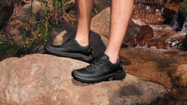 Two feet in black outdoor shoes (Wool Trail X by Giesswein) stand on a brown rock on the bank of a small stream with flowing water.