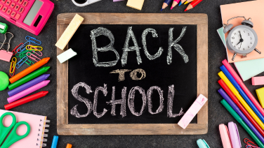 A black slate board with “BACK TO SCHOOL” written in white chalk lies in the centre on a dark background. Various school supplies are arranged around it.