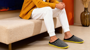 Person sitting on a beige sofa wearing dark grey Giesswein wool slippers with yellow trim, white trousers and a mustard yellow jumper in a modern living room.