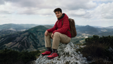 A man in a red winter jacket sits on a rock in the mountains. He is wearing beige trousers, red hiking boots and a rucksack. In the background, you can see wooded hills and a cloudy sky.