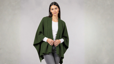 Woman wearing dark green Giesswein merino cape over white top and grey trousers against light grey background