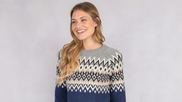 Smiling woman with long blonde hair wearing a grey Giesswein jumper with a navy blue Norwegian knit pattern against a light background