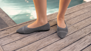 Feet clad in grey knitted ballet flats, resting on a wooden walkway by the edge of a swimming pool.