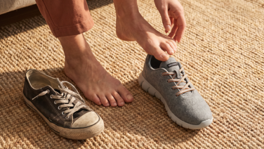 A person is putting on a pair of grey Giesswein Merino trainers on a sisal rug; next to them is a worn-out old shoe.