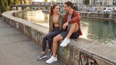 A couple are sitting on a wall by a river in the city, laughing; they’re both wearing grey Giesswein Merino Runner trainers