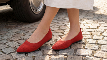A close-up of feet walking in red Giesswein knitted ballerina flats with a textured striped pattern, on cobblestones.