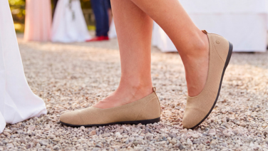 A close-up of feet wearing beige Giesswein knitted ballerina flats on a pebble path, with a festively laid table in the background at a celebration.