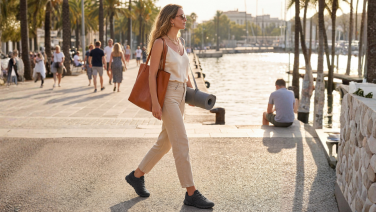 A woman with a bag and a yoga mat is walking along a beach promenade lined with palm trees, wearing black Giesswein Wooltastic trainers.