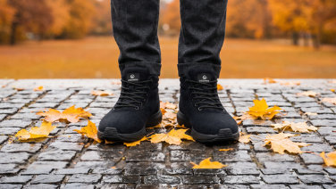 Front view of black, ankle-high Giesswein Merino trainers with laces and the brand logo on cobblestones, surrounded by yellow maple leaves.