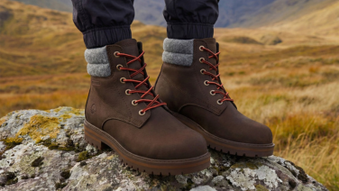 A close-up of the feet of a person standing on a rock in an autumnal mountain landscape. They are wearing brown leather boots with red laces, a grey woollen felt collar and a treaded sole.