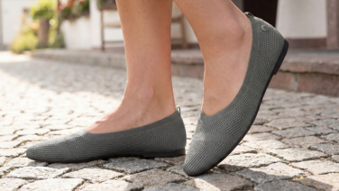 A close-up of feet in grey Giesswein knitted ballerina flats, standing on cobblestones.