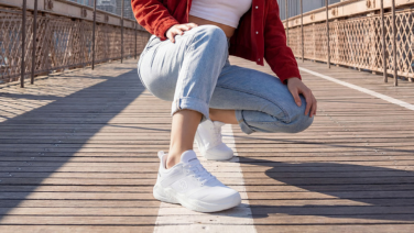 A close-up of a person’s legs in white Giesswein trainers with a textured sole on a sun-drenched wooden walkway. The Giesswein logo is visible on the side of the shoe.
