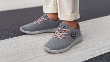 Close-up of a person's feet in cream-coloured, rolled-up trousers. They are wearing grey Merino wool sneakers with apricot-coloured laces and a light grey sole. The person is standing on a zebra crossing.