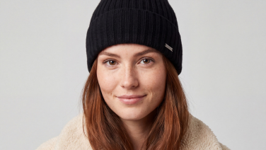 A portrait of a smiling woman with reddish-brown hair and a black ribbed knitted hat with a metal label; she is wearing a cream-coloured teddy fleece jacket against a light grey background.