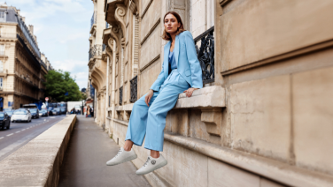 A woman in a light blue trouser suit and white merino wool trainers sits relaxed on a stone wall in an urban street.