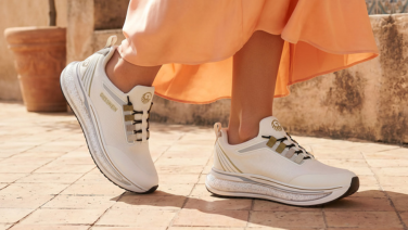 A close-up of a woman’s legs in an orange skirt, standing on cobblestones in the sunlight, wearing white Giesswein trainers with gold detailing and a textured sole.