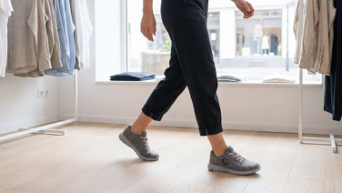 A person wearing dark, rolled-up trousers is walking through a brightly lit shop with a wooden floor; in the background, items of clothing hang on racks. They are wearing grey merino wool lace-up trainers.